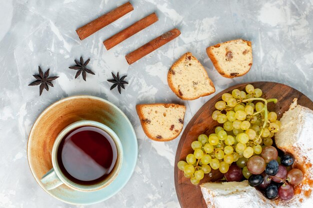 Draufsicht pulverisierter Kuchen köstlicher gebackener Kuchen mit frischen Trauben und Tee auf hellweißer Oberfläche