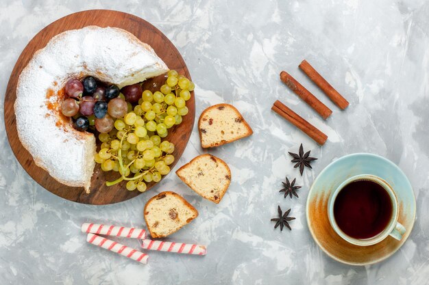 Draufsicht pulverisierter Kuchen köstlicher gebackener Kuchen mit frischen Trauben und Tee auf weißer Oberfläche