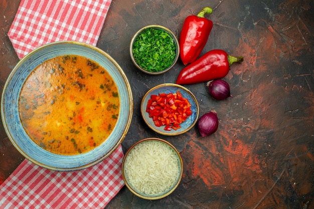 Draufsicht Reissuppe in Schüssel auf rot-weiß karierter Tischdecke rote Paprika Zwiebeln Petersilie andere Sachen in Schüsseln auf Hintergrund