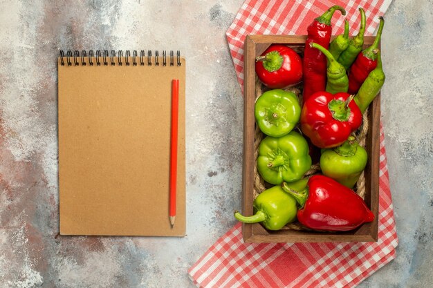 Draufsicht rote und grüne Paprikaschoten Peperoni in der Holzkiste auf kariertem Tischtuch-Notizbuch-Rotstift auf nackter Oberfläche