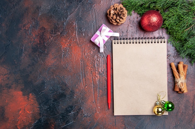 Draufsicht roter Stift ein Notizbuch Kiefer Zweige Weihnachtsbaumball Spielzeug Zimtstangen auf dunkelroter Oberfläche Freiraum Weihnachtsfoto