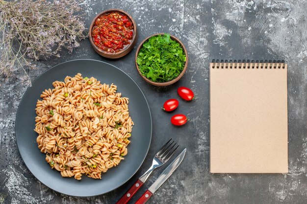 Draufsicht Rotini Nudeln auf runder Platte Tomatensauce gehackte Petersilie in kleinen Schalen Kirschtomaten Gabel und Messer Notizbuch auf dunklem Tisch