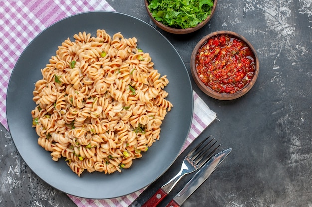 Draufsicht Rotini Nudeln auf Teller auf rosa weiß karierten Serviettengabel und Messer gehacktes Grün und Tomatensauce in Schalen auf grauem Tisch