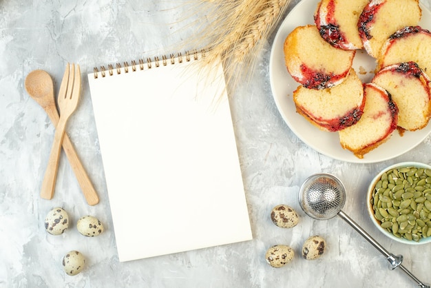 Kostenloses Foto draufsicht scheiben marmeladenkuchen auf teller kürbiskerne in schüssel weizenspitzen notizblock gekreuzte holzgabel und löffel wachteleier auf dem tisch