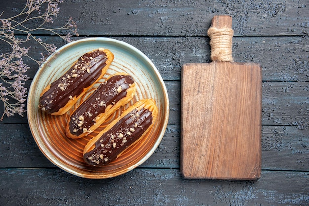 Draufsicht Schokoladen-Eclairs auf ovalem Teller getrockneten Blumenzweig und Schneidebrett auf dem dunklen Holztisch