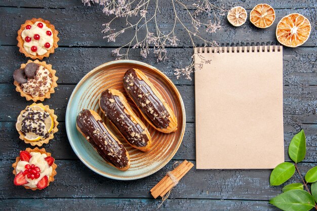 Draufsicht Schokoladen-Eclairs auf ovalem Teller getrockneter Blumenzweig zimtgetrocknete Orangen hinterlässt ein Notizbuch und vertikale Reihenkuchen auf dem dunklen Holztisch