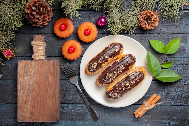 Kostenloses Foto draufsicht schokoladen-eclairs auf weißem ovalem teller tannenbaumzweigen und zapfenweihnachtsspielzeug eine gabel zimt eine tasse tee und ein schneidebrett auf dunklem holztisch