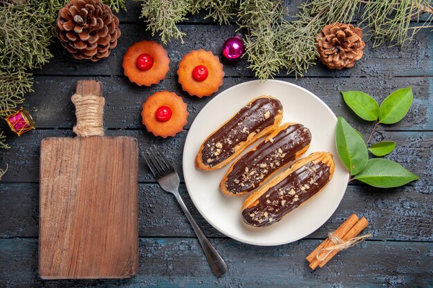 Draufsicht Schokoladen-Eclairs auf weißem ovalem Teller Tannenbaumzweigen und Zapfenweihnachtsspielzeug eine Gabel Zimt eine Tasse Tee und ein Schneidebrett auf dunklem Holztisch