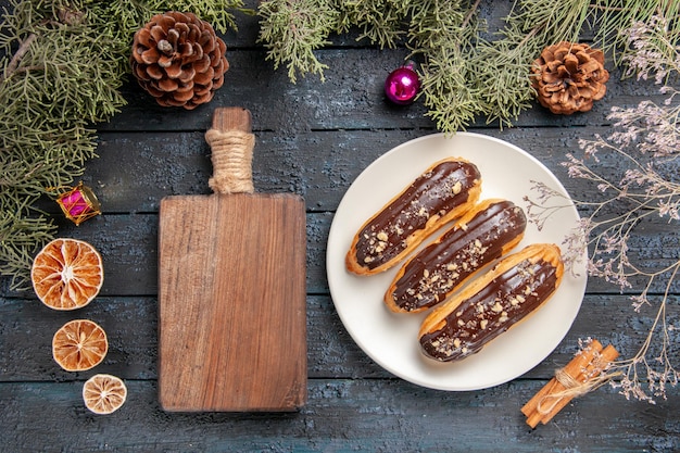 Draufsicht Schokoladen-Eclairs auf weißem ovalem Teller Tannenbaumzweigen und Zapfenweihnachtsspielzeug getrockneter Blumenzweig getrockneter Orangenzimt und Schneidebrett auf dunklem Holztisch