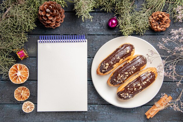 Draufsicht Schokoladen-Eclairs auf weißem ovalem Teller Tannenbaumzweigen und Zapfenweihnachtsspielzeug getrockneter Orangenzimt und ein Notizbuch auf dunklem Holztisch