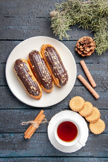 Draufsicht Schokoladen-Eclairs auf weißem ovalem Tellerkegel Tannenbaumblätter Zimt verschiedene Kekse und eine Tasse Tee auf dunklem Holzboden