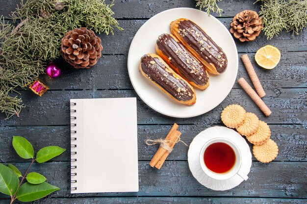 Draufsicht Schokoladen-Eclairs auf weißen ovalen Tellerkegeln Tannenbaumblätter Weihnachtsspielzeug Zimtscheibe Zitrone verschiedene Kekse eine Tasse Tee und ein Notizbuch auf dunklem Holzboden