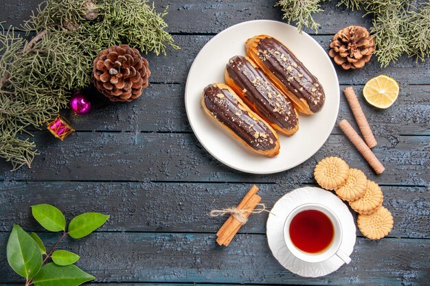 Draufsicht Schokoladen-Eclairs auf weißen ovalen Tellerkegeln Tannenbaumblätter Zimtscheibe Zitrone verschiedene Kekse und eine Tasse Tee auf dunklem Holzboden mit Kopierraum