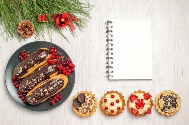 Draufsicht Schokoladen-Eclairs und Johannisbeeren auf dem grauen Teller ein Notizbuch Törtchen am Boden und Kiefernblätter mit Weihnachtsspielzeug auf dem weißen Holzboden