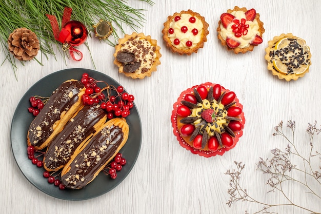 Draufsicht Schokoladen-Eclairs und Johannisbeeren auf dem grauen Teller tört Beerenkuchen und Kiefernblätter mit Weihnachtsspielzeug auf dem weißen Holztisch