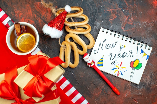 Draufsicht Tasse Tee Mini-Weihnachtsmütze auf roter Tischdecke Holzblöcke ovale Bagels Neujahr auf Notizblock-Geschenken auf dunkelrotem Tisch geschrieben