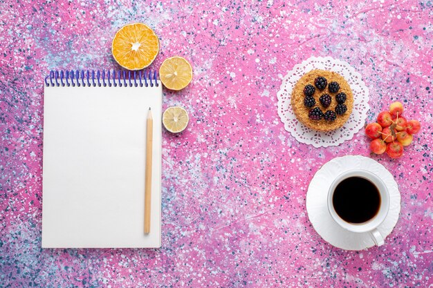 Draufsicht Tasse Tee mit kleinem Kuchen und Notizblock auf dem rosa Hintergrund.