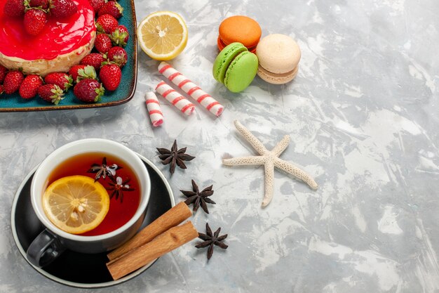 Draufsicht Tasse Tee mit Macarons und kleinem Erdbeerkuchen auf hellweißer Oberfläche