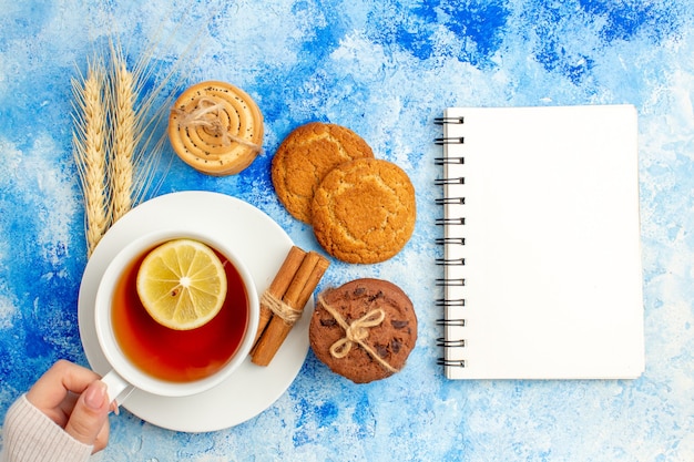 Draufsicht Tasse Tee mit Zitrone und Zimt im weiblichen Handnotizblock auf blauem Tisch gewürzt