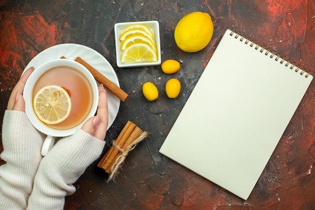 Draufsicht tasse tee mit zitronengeschmack in frauenhänden cumcuat zitronenscheiben in kleine schüssel gebundene zimtstangen notizbuch auf dunkelrotem tisch