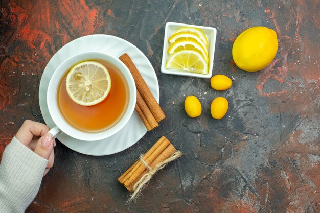 Draufsicht Tasse Tee mit Zitronengeschmack in weiblicher Hand cumcuat Zitronenscheiben in kleiner Schüssel Zimt auf dunkelrotem Tisch