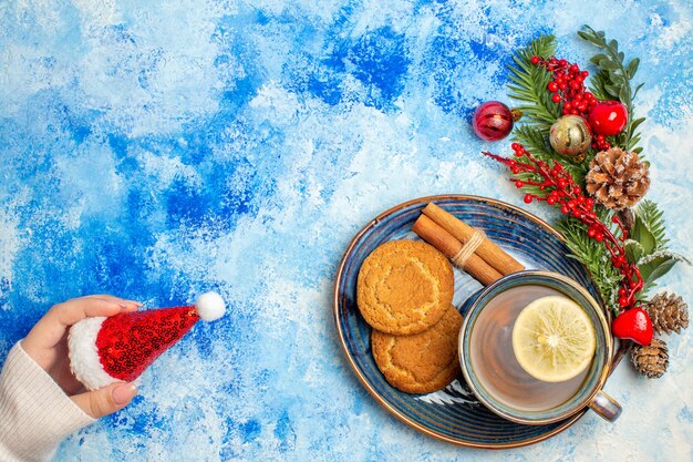 Draufsicht Tasse Tee Zimtstangen Kekse auf Untertasse Weihnachtsmütze auf blauem Tisch Freiraum