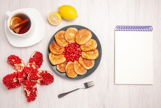 Draufsicht Tee und Pfannkuchen eine Tasse Tee mit Zimt und Zitrone geschälte Granatapfel-Zitrone neben dem weißen Notizbuchteller mit Samen von rotem Granatapfel und Pfannkuchen und Gabel auf weißem Hintergrund