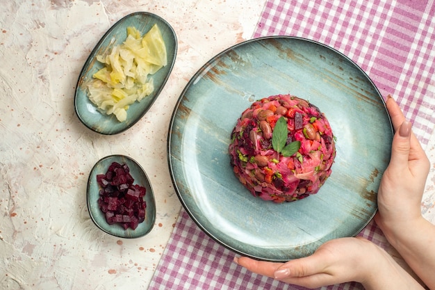 Draufsicht-Vinaigrette-Salat auf ovalem Teller in Frauenhand auf weiß-violett karierten Tischdecken mit einigen Stoffen auf hellgrauem Tisch