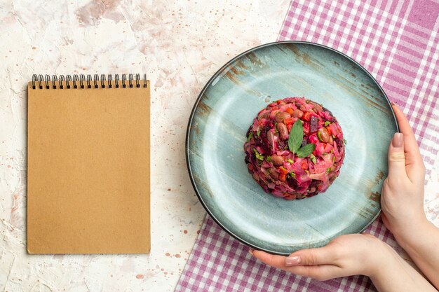 Draufsicht-Vinaigrette-Salat auf ovalem Teller in Frauenhand auf weiß-violett karierter Tischdecke ein Notizbuch auf hellgrauem Tisch