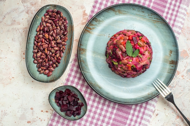 Draufsicht-Vinaigrette-Salat und eine Gabel auf ovalem Teller auf weiß-violett karierten Tischdecken mit Bohnen und geschnittener Rüben auf hellgrauem Tisch