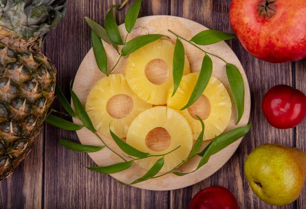 Draufsicht von Ananasscheiben mit Blättern auf Schneidebrett und Ananas-Granatapfel-Pfirsichpflaume auf Holzoberfläche