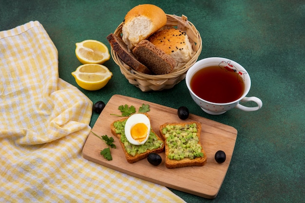 Draufsicht von Avocadopulpen auf geröstetem Brot mit Ei auf hölzernem Küchenbrett mit einer Tasse Tee und einem Eimer Brot auf gre