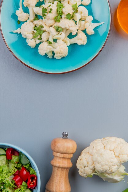 Draufsicht von Blumenkohlstücken mit Koriander in Platte und Salzbutter und Gemüsesalat auf blauem Hintergrund