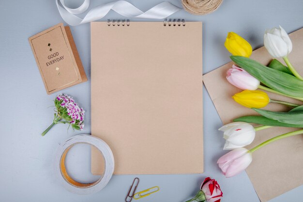 Draufsicht von braunem Blatt Papier mit Postkarte und bunten Tulpenblumen mit türkischer Nelke und Rose auf blauem Hintergrund
