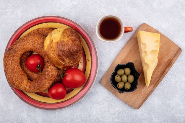 Draufsicht von Brötchen auf einem Teller mit frischen Tomaten mit Oliven auf einer schwarzen Schüssel und Käse auf einem hölzernen Schneidebrett auf einem weißen Hintergrund