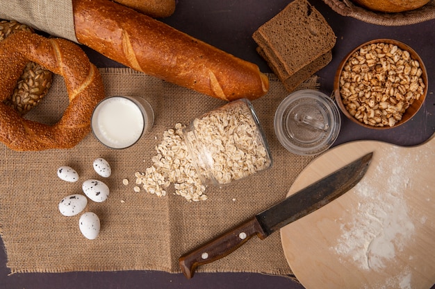 Draufsicht von Broten als Bagelroggen und Baguette mit Milchhaferflocken-Eiern, Körnern und Messer auf Sackleinen auf kastanienbraunem Hintergrund