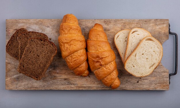 Draufsicht von Broten als Croissant geschnittener Roggen und Baguette auf Schneidebrett auf grauem Hintergrund