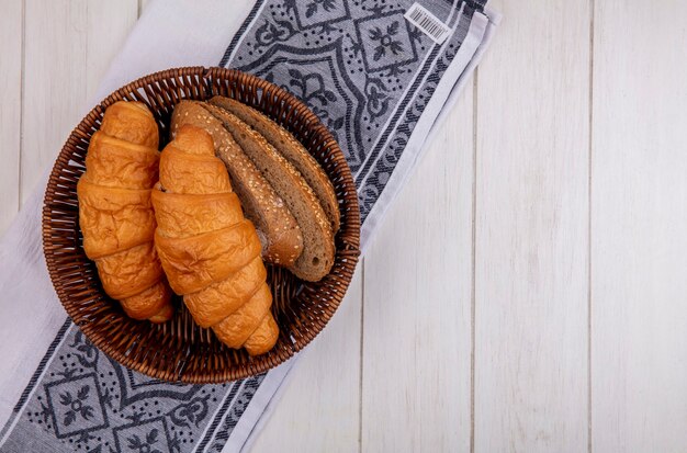 Draufsicht von Broten als Croissant und gesäte braune Kolbenbrotscheiben im Korb auf Stoff auf hölzernem Hintergrund mit Kopienraum