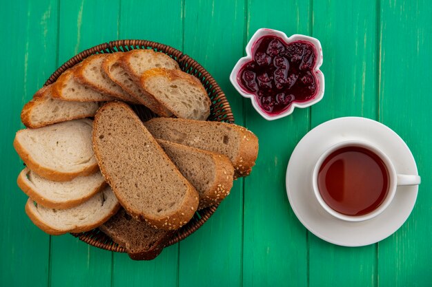 Draufsicht von Broten als gesäte braune Kolben- und Baguettescheiben im Korb und Himbeermarmelade in der Schüssel mit Tasse Tee auf grünem Hintergrund