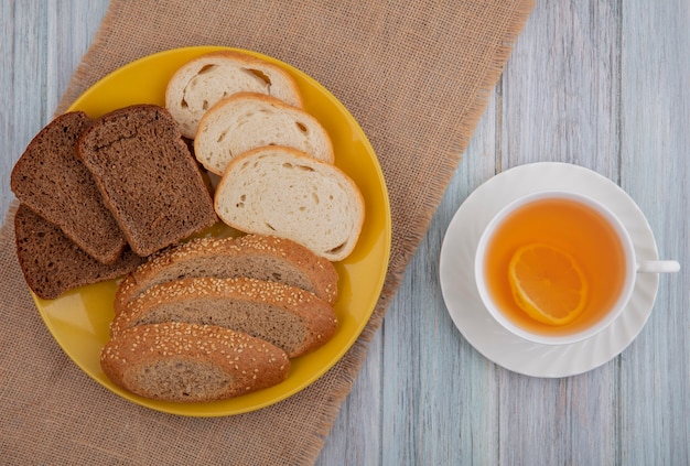 Draufsicht von Broten als geschnittene weiße Samen des braunen Kolbenroggens in Platte auf Sackleinen und Tasse heißen Wirbel auf hölzernem Hintergrund
