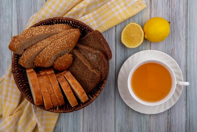 Draufsicht von Broten als geschnittener brauner Kolbenroggen und knusprige im Korb auf kariertem Stoff und Tasse heißem Wirbel mit halb geschnittener Zitrone auf hölzernem Hintergrund