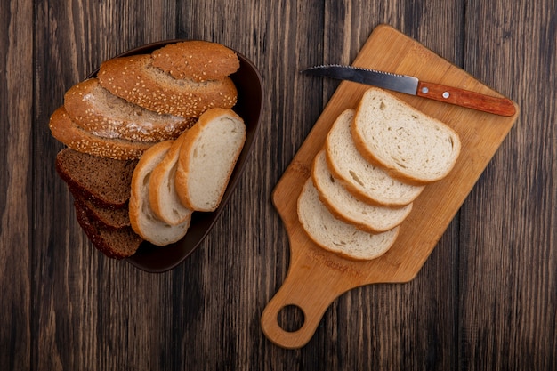 Draufsicht von Broten als geschnittener gesäte brauner Kolbenroggen und weiße in der Schüssel und auf Schneidebrett mit Messer auf hölzernem Hintergrund