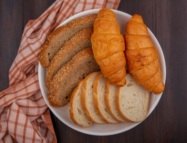 Draufsicht von Broten als geschnittenes Baguette mit braunem Cob und Croissants in Platte auf kariertem Stoff auf Holzhintergrund