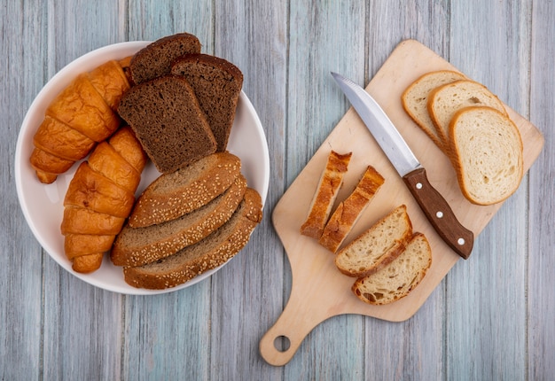 Draufsicht von Broten als geschnittenes Baguette mit Messer auf Schneidebrett und Croissantroggen und gesätem braunem Kolben in Schüssel auf hölzernem Hintergrund