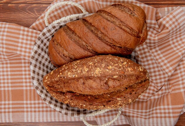 Draufsicht von Broten als schwarzes und entkerntes Baguette im Korb auf kariertem Stoff und Holztisch