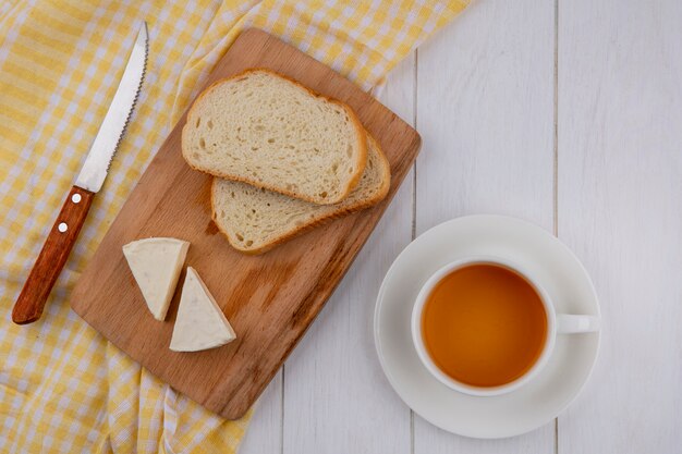 Draufsicht von Brotscheiben mit Käse auf einer Tafel mit einem Messer auf einem gelben Handtuch und einer Tasse Tee auf einer weißen Oberfläche