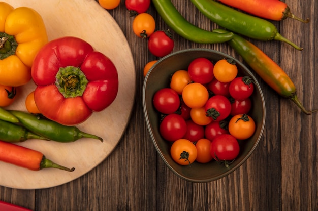 Draufsicht von bunten Paprikaschoten auf einem hölzernen Küchenbrett mit Kirschtomaten auf einer Schüssel auf einer Holzwand