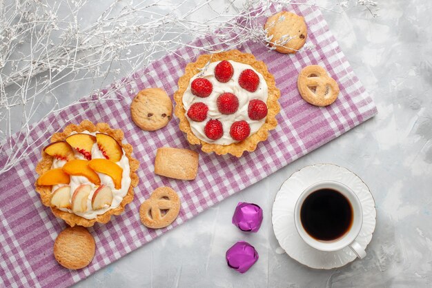 Draufsicht von cremigen Kuchen mit weißer leckerer Sahne und geschnittenen Erdbeerenpfirsichen Aprikosen mit Keksen und Tee auf hellem Schreibtisch, Obstkuchencreme backen