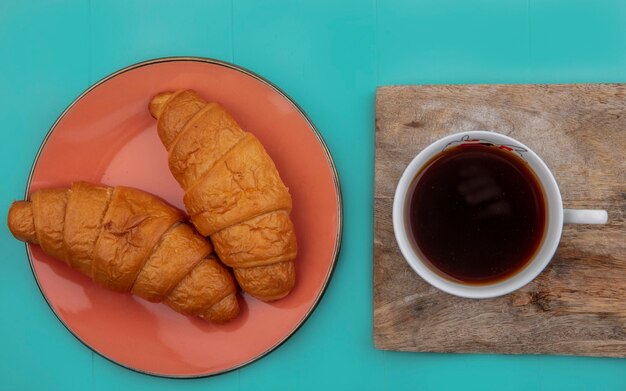 Draufsicht von Croissants in Platte und Tasse Tee auf Schneidebrett auf blauem Hintergrund
