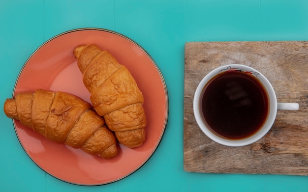 Draufsicht von Croissants in Platte und Tasse Tee auf Schneidebrett auf blauem Hintergrund